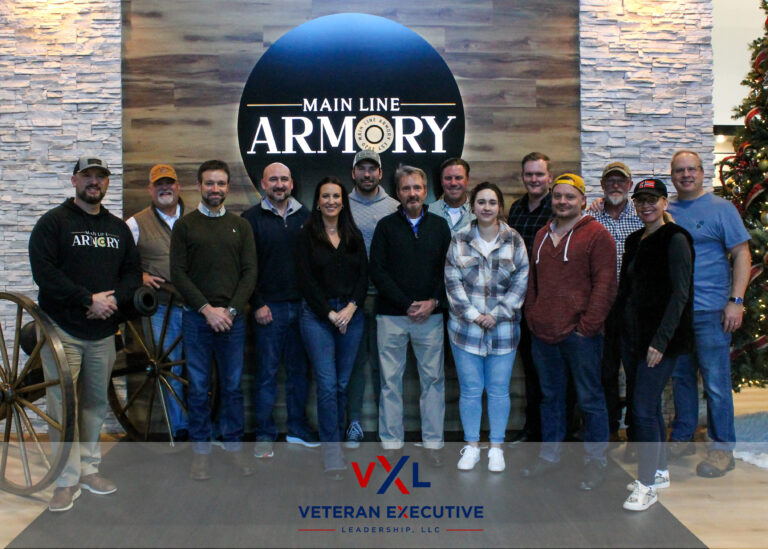 A group of people pose for a photo at an armory. They are standing in front of a sign that reads Main Line Armory.